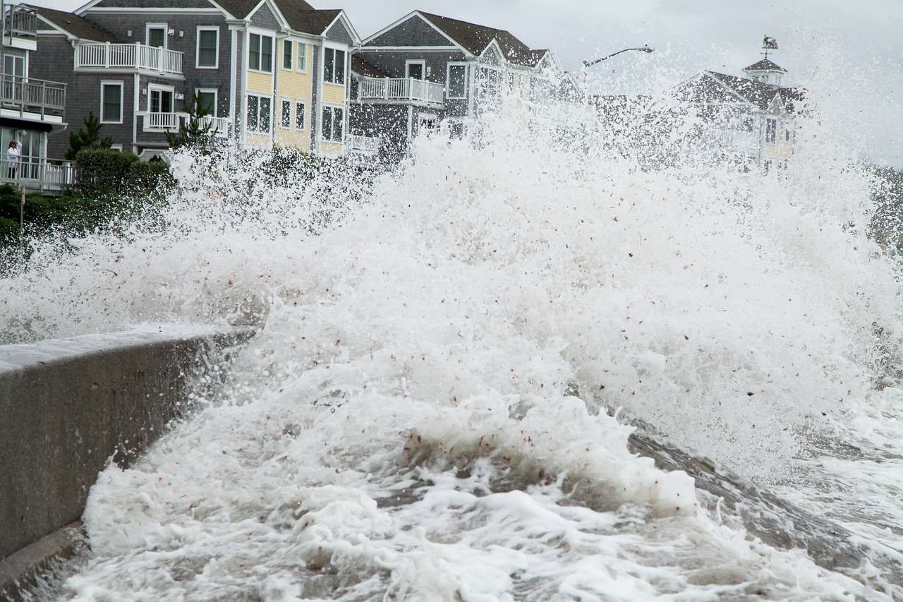 Close-up from the water side, of a wave breaking over a shore barrier in front of houses.