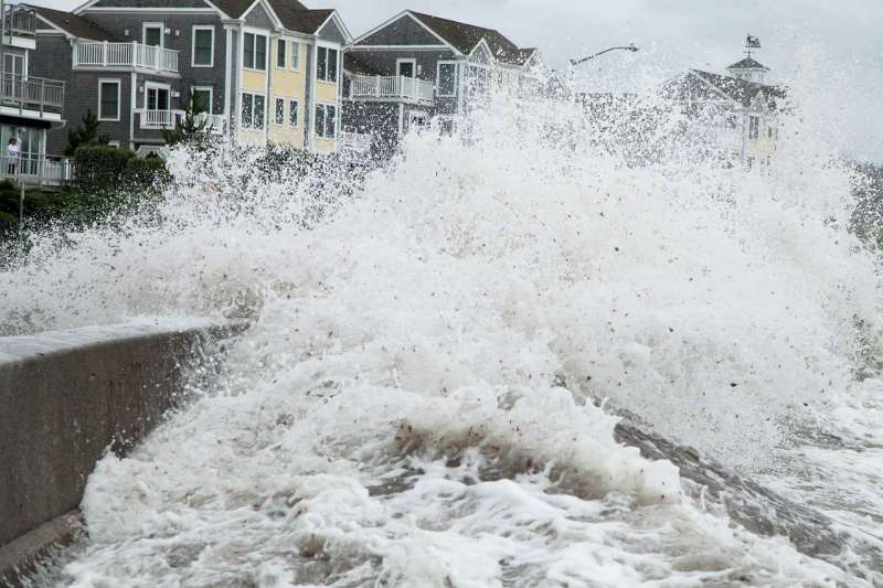 Close-up from the water side, of a wave breaking over a shore barrier in front of houses.