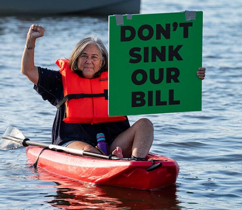 Woman in kayak holding sign, Don't sink our bill.
